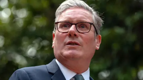PA Media A man with grey hair and dark glasses in s close-up shot. He is wearing a dark suit, white shirt and grey tie. There are trees in the background. 