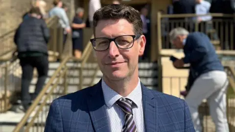 Dane Harrop, who has short brown hair and is wearing dark rimmed glasses and a blue chequered suit and stripy tie. There are stairs to the building's entrance behind him with people walking around.