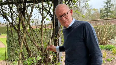 Clare Worden/BBC David Horton-Fawkes stands next to some of the damaged roses. He is wearing a blue jumper and dark-rimmed glasses. 