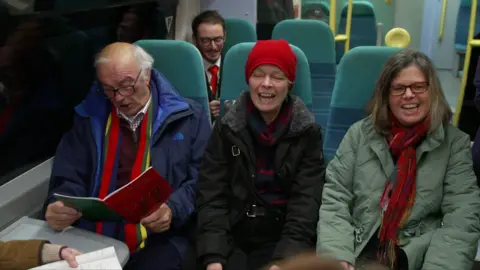 Three people, a man and two women, wearing winter clothing, sit on a Southern train singing from sheet music.