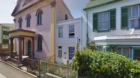 The Jersey Islamic Centre is a former Methodist church and is painted pink with yellow and burgundy trimmings. Next to it are two residential homes, one small pink one and one larger white one with green shutters and a hedge in front of it.
