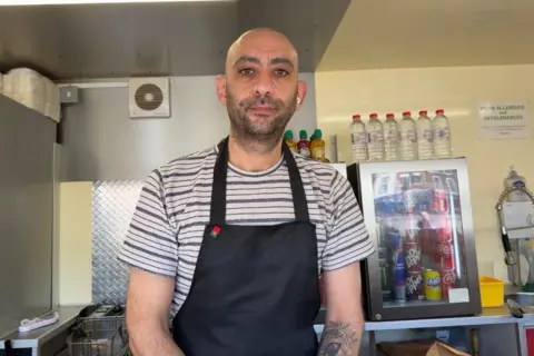 Ollie Conopo/BBC A man in a black and white striped T-shirt and a navy apron stands in a food truck. Behind him are lots of bottles of water and a transparent fridge with soft drinks in.