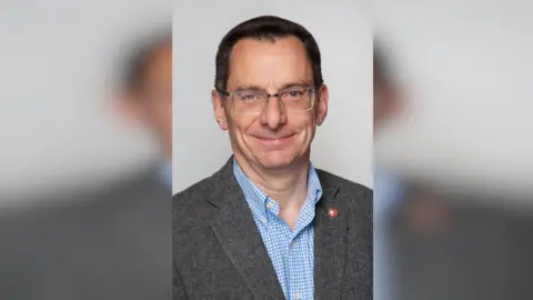 LDRS A man with short black hair in a suit. He is wearing glasses and has a baby blue chequered shirt under his blazer. 