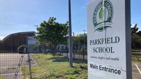 A sign reading 'Parkfield School main entrance' with a sports court and a school building in the background