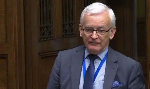 A bespectacled Martin Vickers wearing a suit and tie, with handkerchief in top pocket addressing the Commons