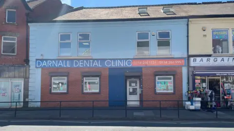 Simon Thake A brick building with a large blue sign stands next to a convenience store with items spilling out over the pavement