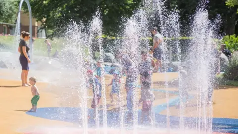Children and parents play under water fountains at a park play area. There is a yellow and blue floor with water fountains dotted across it. There are lawns and trees in the background, as well as some houses that are partly visible on the edge of the park.