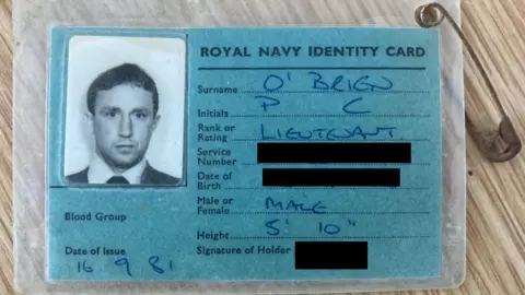 Royal Navy The identity card has a head and shoulders shot in black and white photo of a man wearing a white shirt, dark jumper and tie. It is labelled ROYAL NAVY IDENTITY CARD and in biro is written: O'Brien, PC, Lieutenant, Date of Issue 16.9.81, Height 5'10". The date of birth and the service number is redacted. The card is encased in plastic and has a safety pin on the top right hand corner.