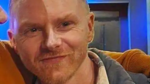 A man with a light beard smiles while another person, who is out of shot, has his arm over his shoulder.