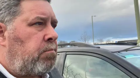 A profile picture of Malloy next to his car. He grey hair and a grey/white beard.