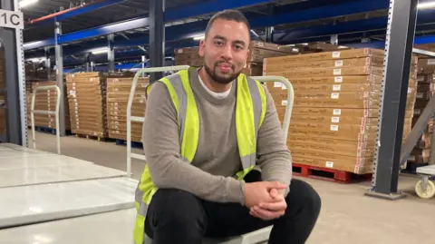 Dan Beckles wearing a high-vis jacket sitting on a trolley in a warehouse with stacks of boxes behind him.