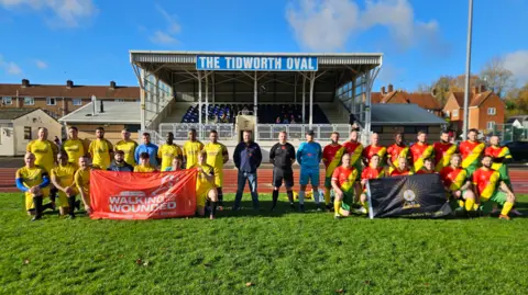 Walking With The Wounded Men in football kits on a football pitch holding flags which say 'Walking for Wounded' A stand is behind them which reads 'The Tidworth Oval'
