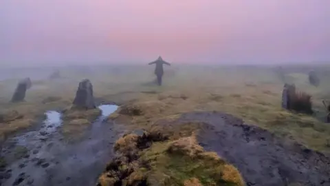 Bunty May Marshall A person stands with arms outstretched among 12 stones in a circle. The stones are about a foot high. There is a pinky-purple sky and it is damp on the moor.
