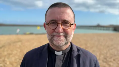 Charlie Rose/BBC Standing on a shingle beach in the evening sunshine, Archdeacon Martin Lloyd Williams, with glasses and short cropped hair, looks at the camera.