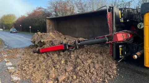 Peter Sutton A yellow and red lorry overturned close to a junction. It has spilled dirt in the middle of the road.