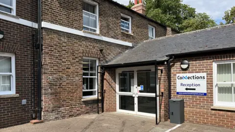 Clare Worden/BBC External view of Connections at St James. It is a two storey brick building with a large white windowed entrance. There is a sign saying "Connections" and an arrow pointing to the door. 