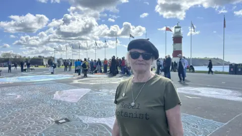 Elizabeth Richmond smiling at the camera in front of a chalk artwork on the promenade which is made up of blue swirls. She has a green T-shirt on which reads 'Rebel Botanist' and is wearing dark sunglasses and a black beret. There is a crowd of people behind her.