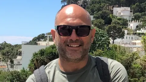 Anthony Barnes is wearing black framed sunglasses, a green T-shirt and a grey backpack. He is standing on a hill which has several white houses and a variation of trees.