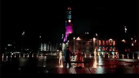 A scene from Virdee with the two protagonists in wedding outfits in front of the mirror pool and City Hall at night