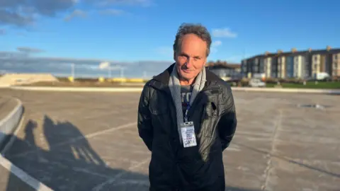 Dale Patterson at the empty lake. He has a short grey hair and is wearing a black jacket and a grey cardigan. His arms are behind his back and he is smiling slightly at the camera. 