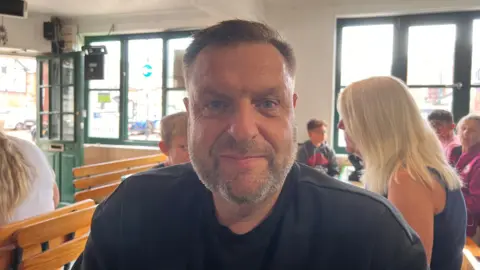 A man in his late 40s with a stubble beard and slicked-back hair, wearing a black T-shirt, looks directly into the can