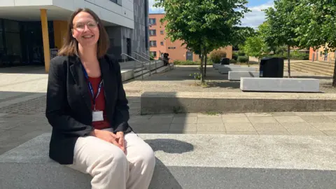 Amy Holmes/BBC A picture of a woman sat down outside in the sunshine. She is wearing white trousers, a black jacket and a red top. She has glasses on and shoulder length brown hair. There are trees and buildings behind her.