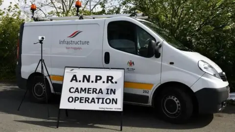 DOI A white van parked on a road near some green trees. It has a DOI logo and a yellow stripe on the side and there is a camera on a tripod in front of it. Next to it there is a white sign with black writing that reads ANPR camera in operation.