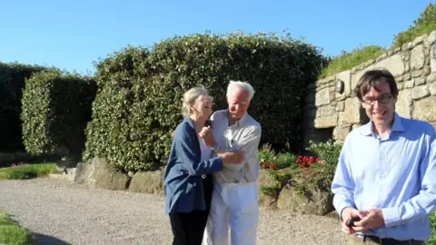 PA Media A man and a woman laughing with their arms round each other, standing on a gravel driveway with a wall and a large bush behind them. A man stands on the right hand side, laughing and looking at the camera. It's a sunny day and the sky is blue.