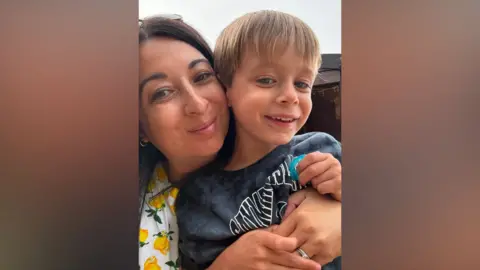Submitted Natalie, who has dark, centre-parted hair and glasses on top of her head, is wearing a white and yellow patterned top. She is holding Freddie, who has fair hair brushed forward. He is wearing a grey T-shirt with writing across the front. Both are looking at the camera and smiling.