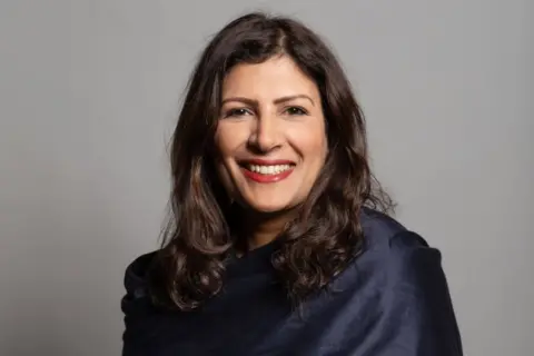 A woman with long dark hair smiles into the camera. She has red lipstick on and is wearing a dark blue top