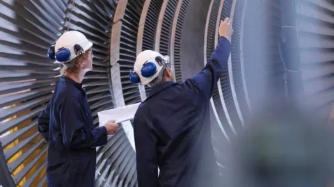 Two men wearing navy blue overalls, white hard hats with blue headphones on. They are stood in front of metal machinery.