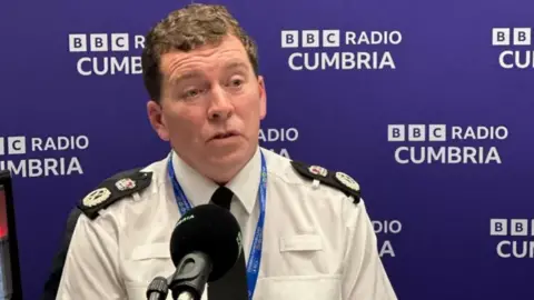 A wide shot of Rob Carden, in his police uniform. He has curly brown hair and is sitting in front of a black microphone to be interviewed for BBC Radio Cumbria.