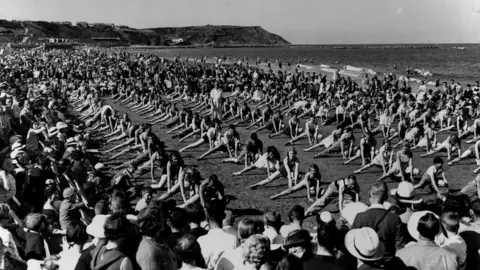 North Yorkshire Archives The image shows hundreds of young women all wearing similar swimsuits performing synchronised stretching or calisthenics on a beach.
They are arranged in neat, evenly spaced rows.