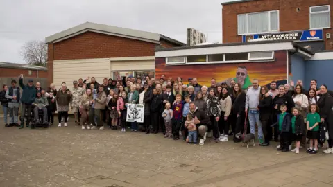 About 80 people stood in front of a mural of a man sat on rocks, with the sunset behind