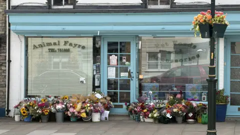 PA Media Dozens of bouquets of flowers sit in vases on the floor outside a pet shop. The storefront is traditional, it is painted light blue and has blinds down in its window. Lettering on the window reads 'Animal Fayre'.