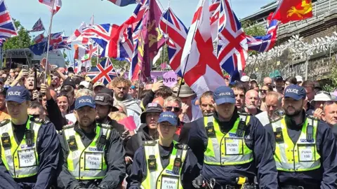 Police officers standing in front of a group of protesters who are holding a variety of flags, including the Union Jack and the flag of St George.