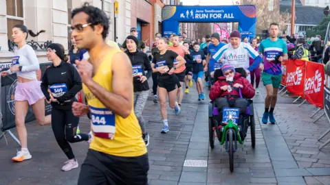 Run for All Runners one in yellow and man pushing a three-wheeled bicycle with Run for All banner in background 