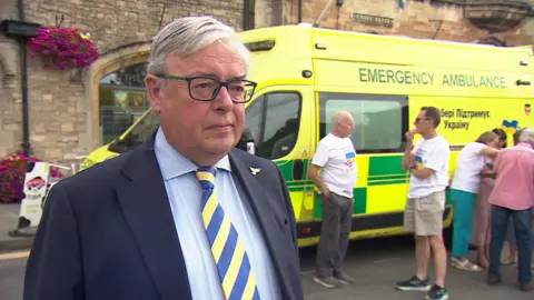 Gavin Grant, who is wearing a suit jacket and shirt with a tie in the same blue and white colours as the Ukraine flag, stands in front of an ambulance.