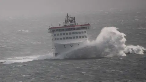 IOMSPC A large wave breaking against the bow of the Manxman ferry in rough seas. It is a large white red and black ferry and the sea surrounding it is grey with white-crested peaks.