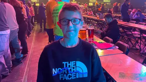 Family A boy with short hair and glasses, with a dark jumper which says The North Face, stands and smiles. Behind him are people standing up and sitting down at lines of tables in a hall.