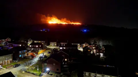 Picture of an orange glowing grassfire in the dark sky, which can be seen on the mountains behind a residential area. Several houses can be seen below, with their windows lit up. Street lights illuminate the roads, where cars can be seen parked on pavements. One car can be seen driving down a street with headlights on. 