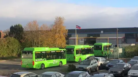 Hereford City Council Three green buses can be seen parked up in a car park, with other vehicles nearby. There are trees and an industrial building in the background.