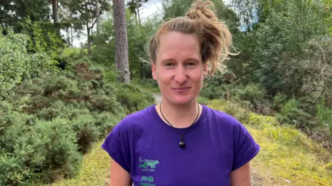 Katie Jachacy is standing outside in an area of woodland. She has her brown hair tied up and is wearing a purple T-shirt.