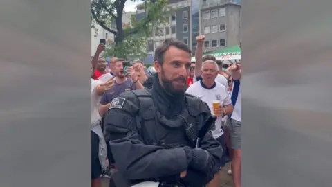 A German police officer with a strong resemblance to Gareth Southgate, surrounded by England fans in a public square in Dortmund.