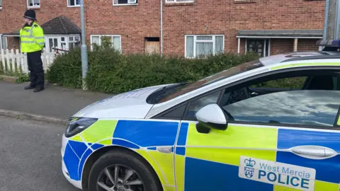 A police officer in a high-vis jacket and police helmet stands outside a row of brick houses. A car labelled "West Mercia Police" is parked nearby.