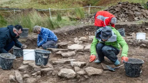 Airborne Lense/Destination Tweed Four people kneeling down on exposed soil and stones with buckets, spades and trowels as they assist with an excavation 