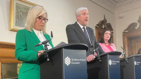 PA Media Northern Ireland First Minister Michelle O'Neill, Infrastructure Minister John O'Dowd and deputy First Minister Emma Little-Pengelly speaking at a press conference at Stormont Castle