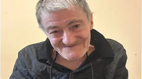 A head shot of an elderly man with grey hair and stubble, wearing a black quilted jacket.