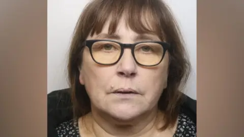 A middle-aged woman with shoulder length brown hair and a fringe. She is wearing black-framed eye glases and posing for a police mug shot.
