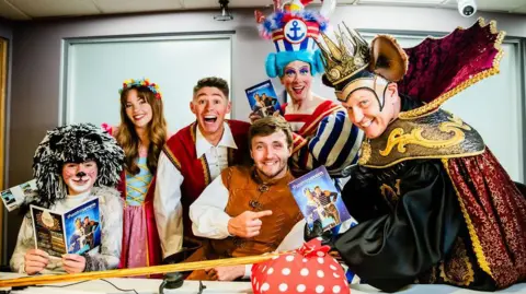 Victoria La Bouchardiere Six people dressed up as pantomime characters smile as they gather around a table in an office to pose for the camera, with some holding the programmes for the pantomime.